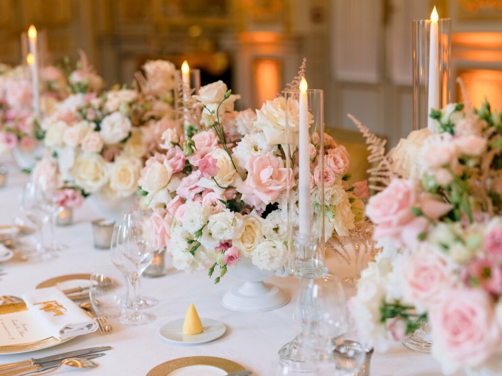 Centres de table de roses poudrées, lisianthus blancs et astilbes dans des coupes blanches, bougies sous photophores et assiettes dorées